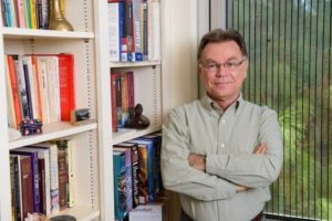 Man with arms folded standing next to bookshelf