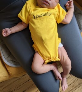 Newborn in a yellow onesie printed with "kindbaby" across the chest