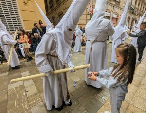 Hooded penitent dripping wax on a child's wax ball during a pause in a Holy Week procession in Malaga, Spain