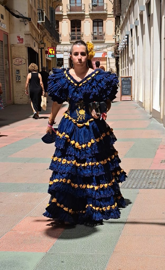 Woman in blue, gold-trimmed Flamenco dress walking down a street in Malaga
