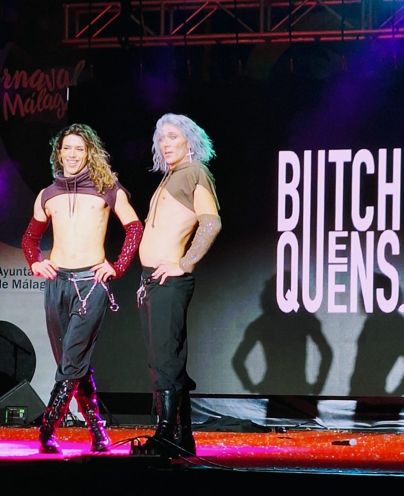 Two men in jeans and exposed chests next to a sign that reads ButchQueens