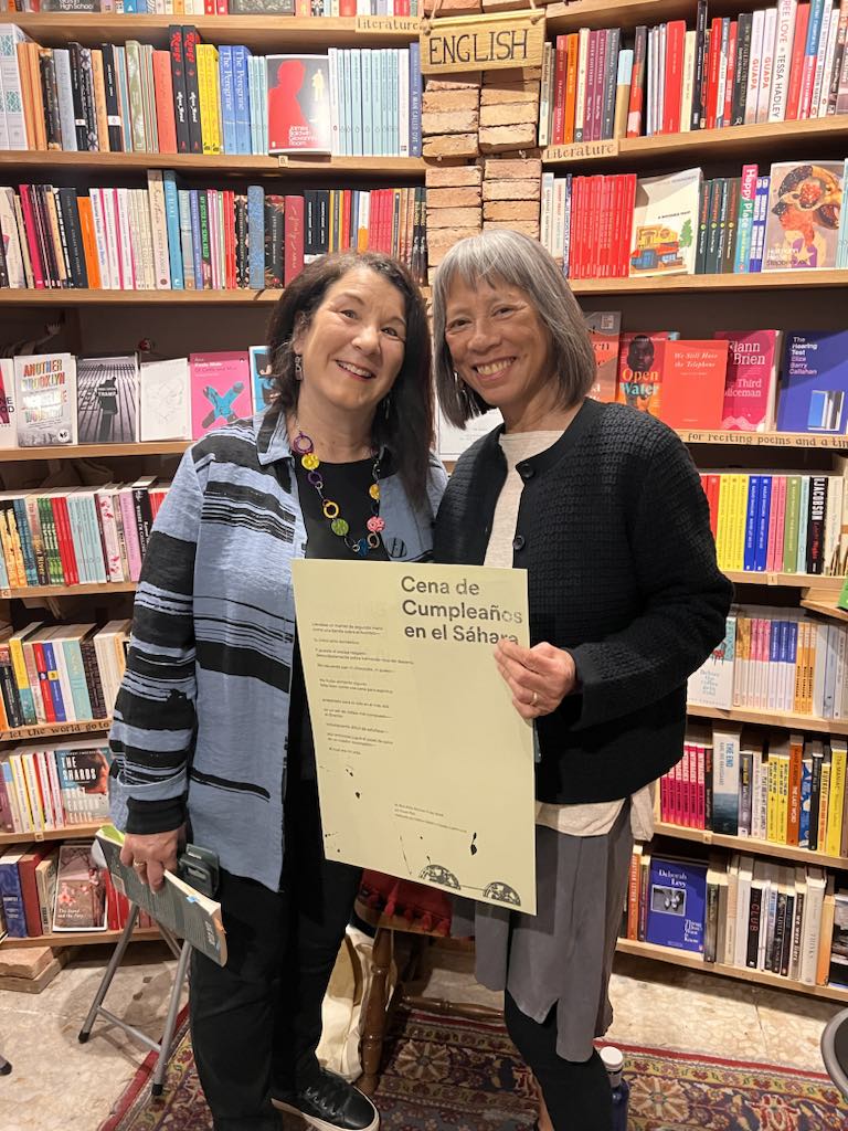 Two women standing in a bookstore, one holding a large sheet of paper with a poem in Spanish printed on it