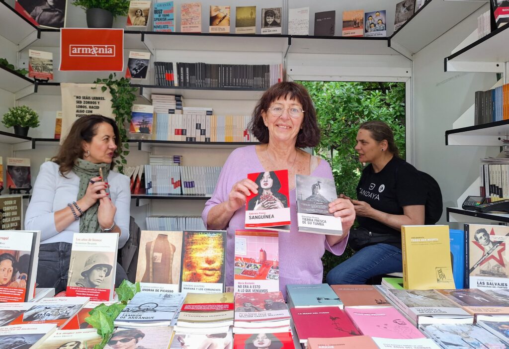 Three women in a book fair booth, the middle one holding up two books for display