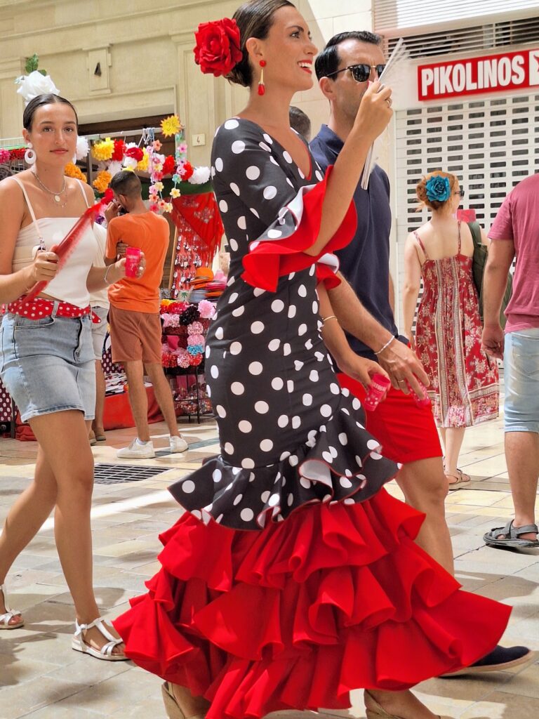 A tall woman in a black and white polka-dot Flamenco dress and red flower in her hair fans herself as she walks