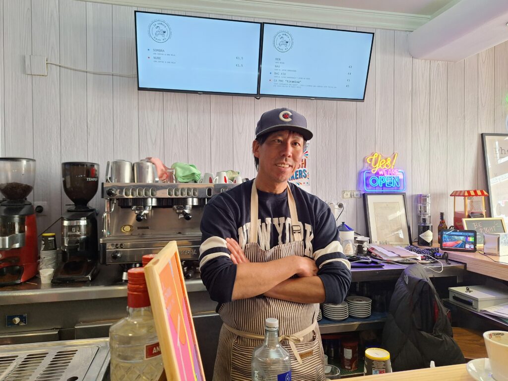 Vietnamese American man in a Chicago Cubs baseball cap stands with arms folded behind the counter of a cafe/bar.