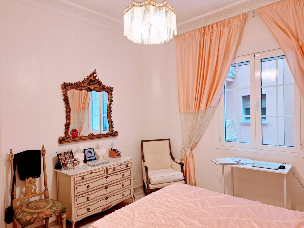 View of a bedroom showing a French style dresser, mirror and two chairs, a chandelier, and pink curtains.