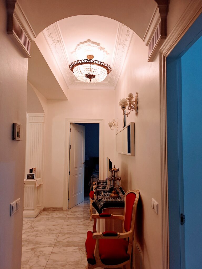 Partial view of the interior of an apartment from the entryway showing a chandelier and sconces and a red Louis XV chair.