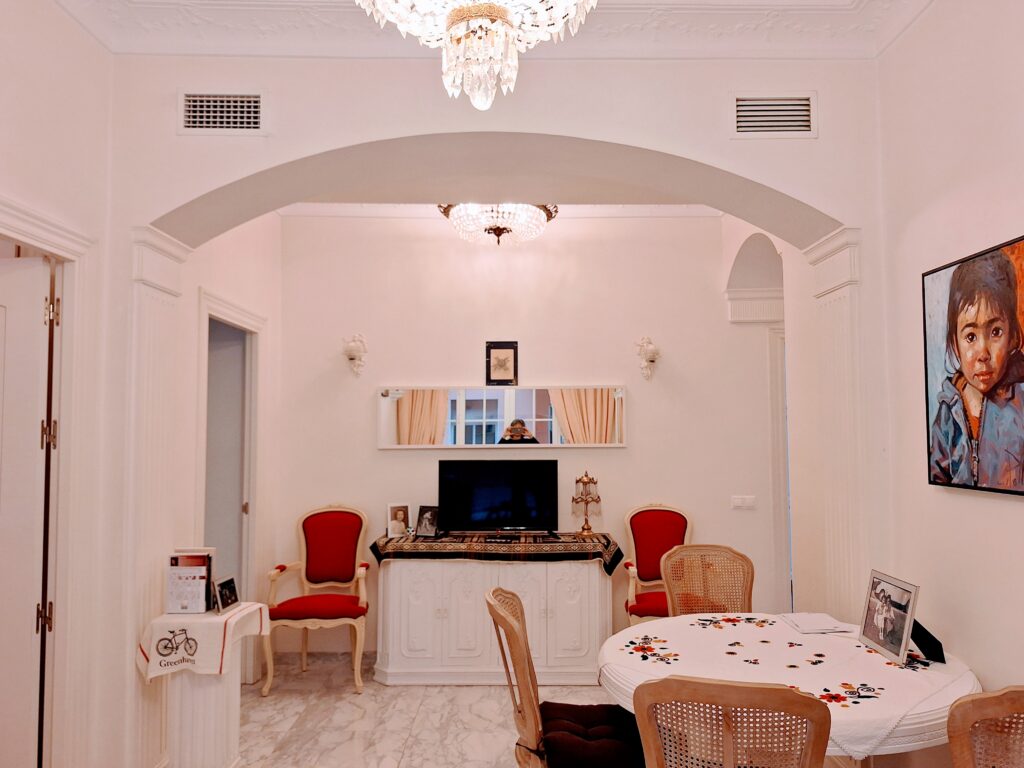 Living/dining area with red Louis XV chairs in the background, a round table with flowered tablecloth in foreground, a portrait painting of a child on the wall on the right, and a chandelier in the ceiling.