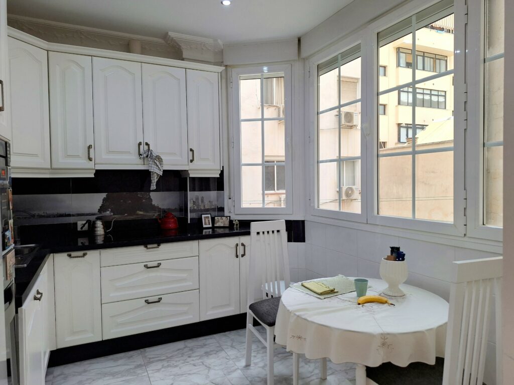Kitchen with white cupboards and black countertop, round table with white tablecloth, white stone floor, and a back of windows that give a view of the adjoining and opposite buildings.