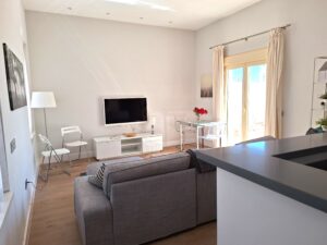 Interior of an apartment with white walls, white TV stand, white lamp and white folding chairs, and gray couch. Glass doors open to a terrace.