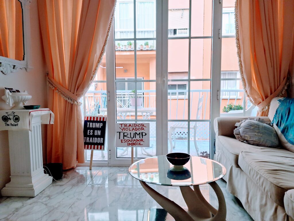 Living room with sofa and small round, glass-top table with a ceramic bowl on top. Against the terrace doors framed by pink curtains are two protest signs.