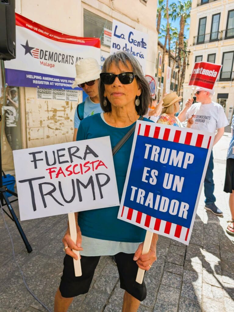 Woman in sunglasses and grim expression holding a protest sign in each hand.