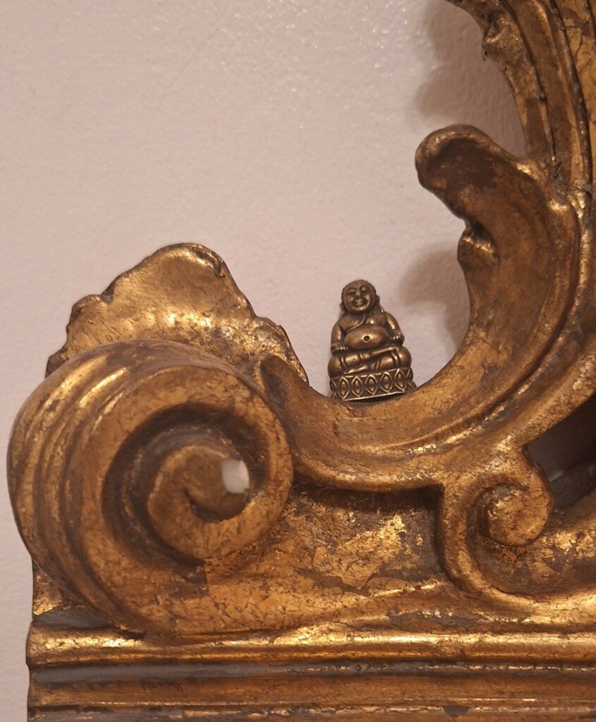 Detail of a gilt mirror showing a tiny figurine of a Buddha perched near the base.