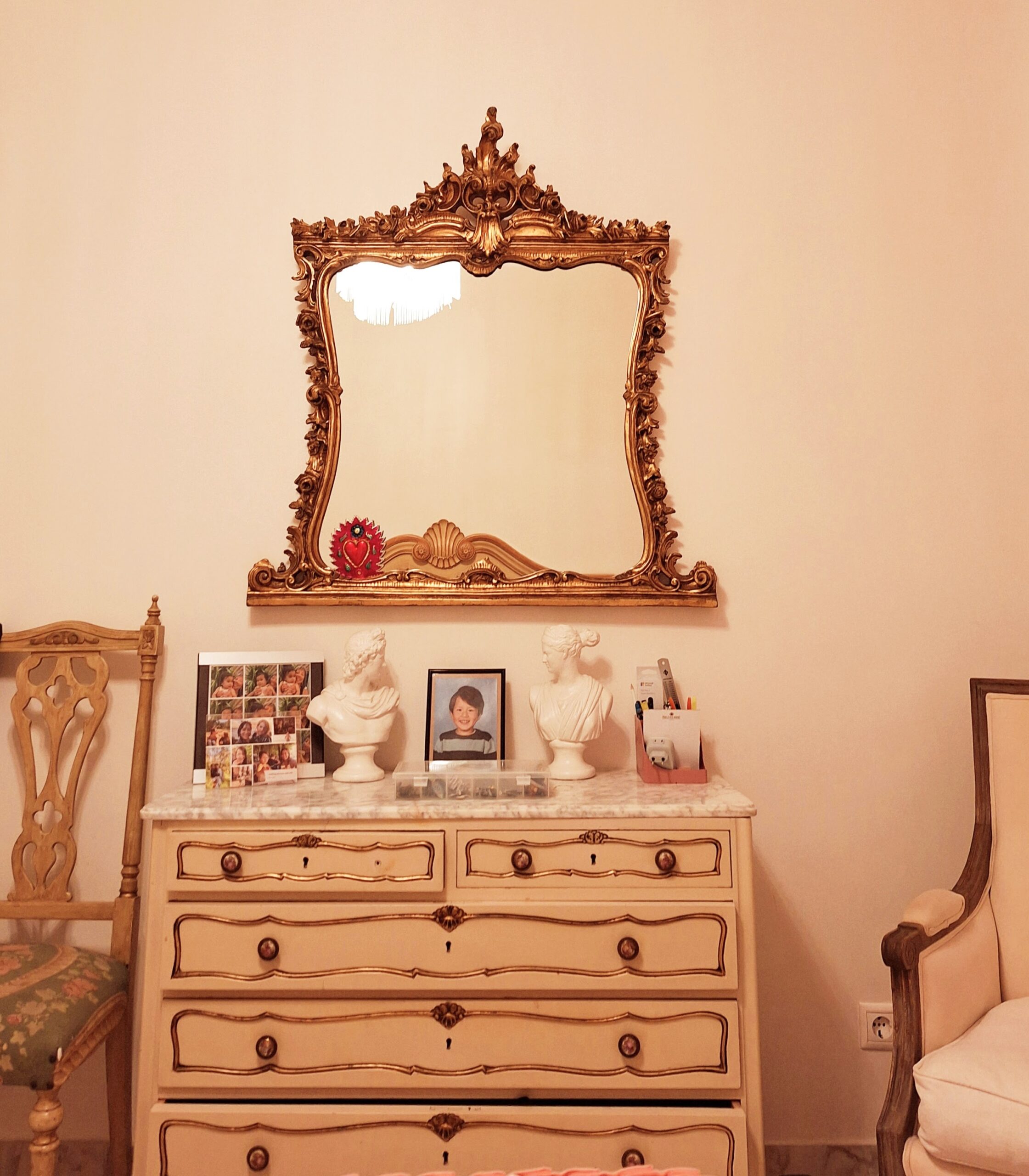 Gilt mirror over a chest of drawers and a chair on either side all in a baroque French style.