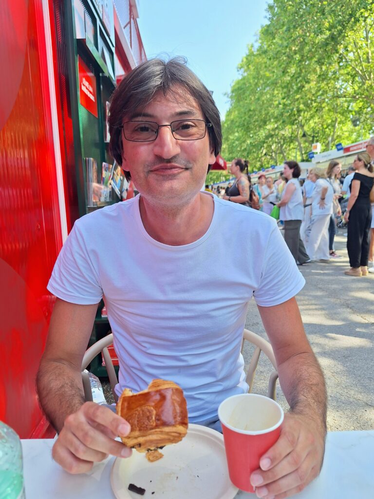 Man with glasses eating a pastry and holding a paper cup at an outdoor fair.