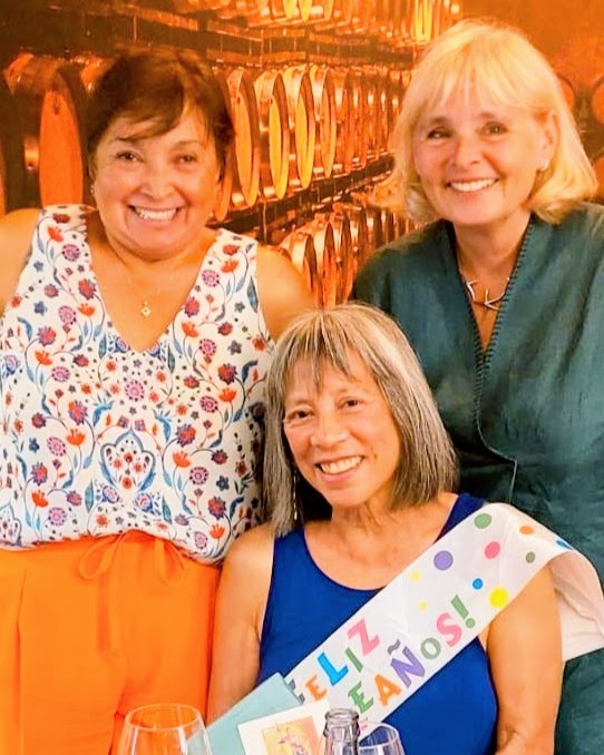 A woman seated at a table with a birthday sash flanked by two women standing.