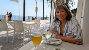 Dark-haired, dark-skinned woman sitting at a seaside cafe with a glass of wine in front of her.