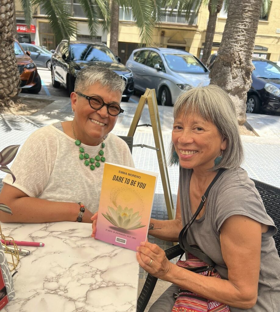 Two brown=skinned women sit at a table holding a book between them.