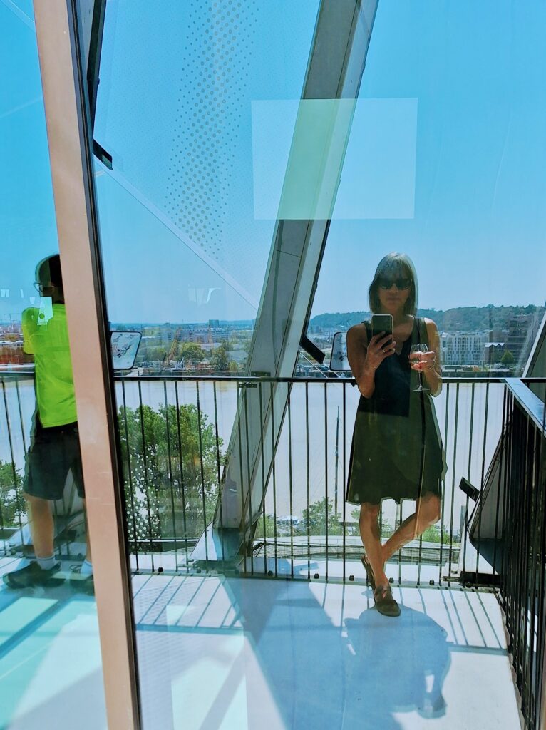 Woman in black dress on a terrace with a view of a river and the city. She's holding a glass of wine and aiming her phone camera at herself.