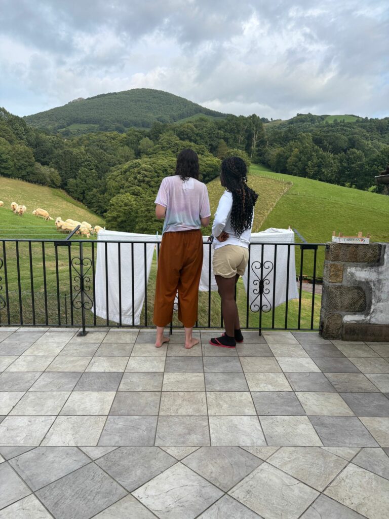 Two tall women look out from a terrace at sheep grazing on the hills.