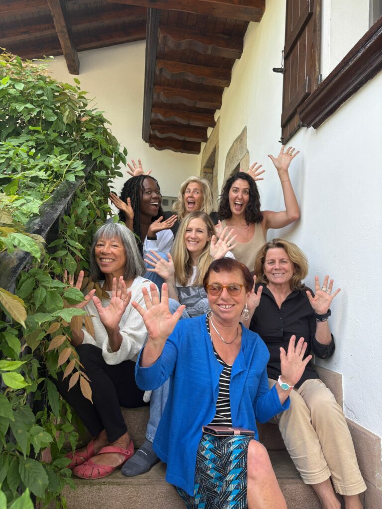 Seven women seated on front steps to a home with both hands raised