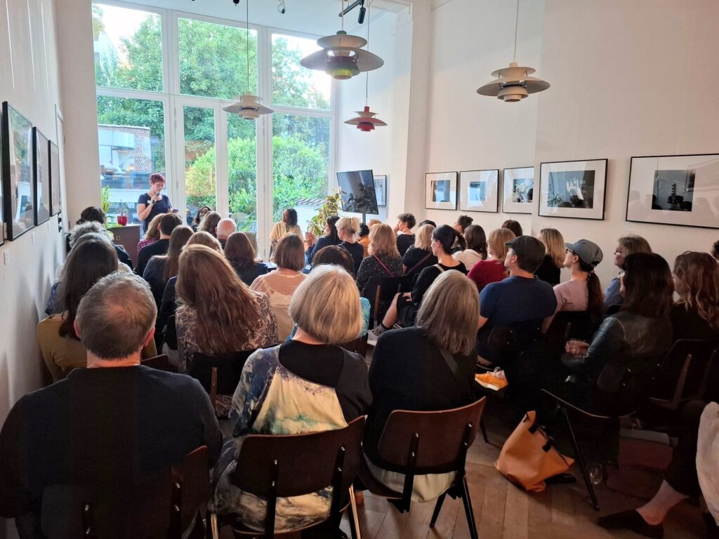 A room full of people facing speakers at the front of the room against a panel of windows looking onto a garden.