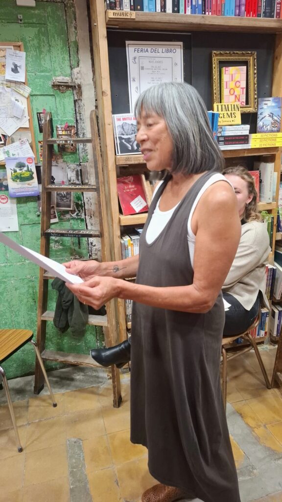 Dark-skinned, gray-haired woman in brown jumper reads from a sheet of paper to an audience in a bookstore.