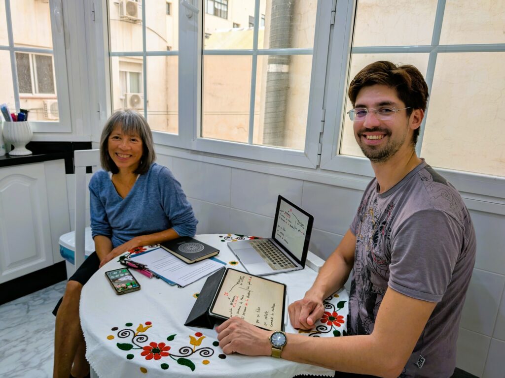 A man and a woman seated at a kitchen table with two laptops for a tutoring session.