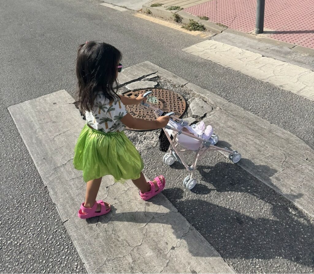 A toddler girl dressed in a neon green skirt and neon pink sandals wears sunglasses as she pushes a stroller with her doll across the street.