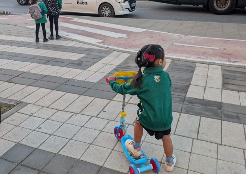 Little girl in green jacket and black shorts with red bow in her hair with one foot on a scooter and the other on the ground