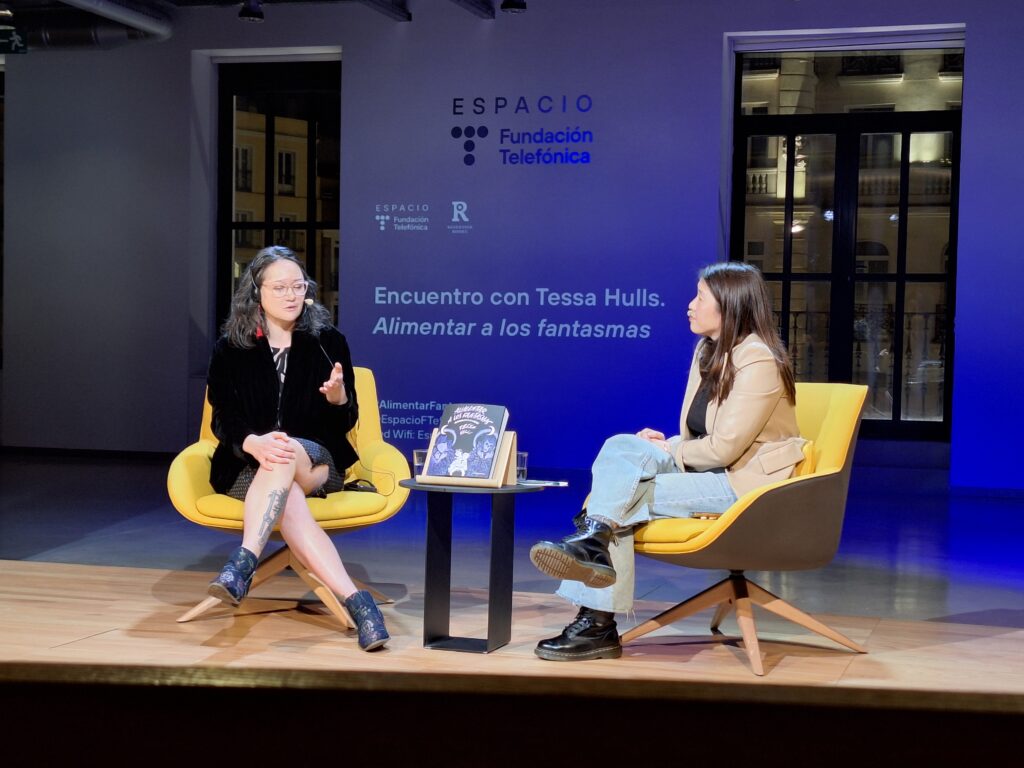 Two women of Asian descent sitting in yellow chairs opposite each other in conversation on stage