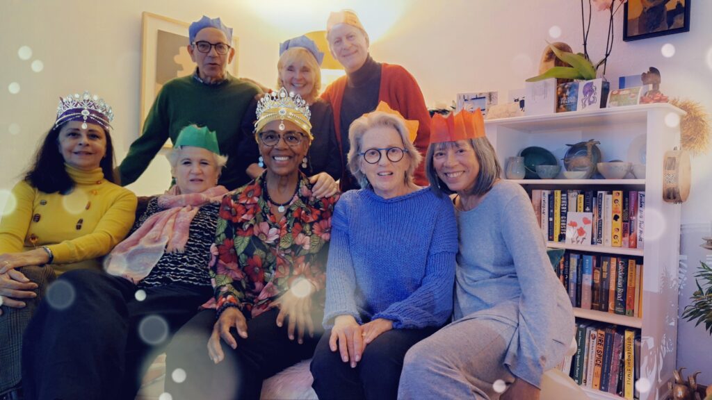 Five women sit on a couch. Behind them stand a woman flanked by two men. Everyone is wearing a paper crown. Light from the lamp in the back causes different sized circles of light to appear like bubbles across the photo and light up two of the crowns like tiaras.