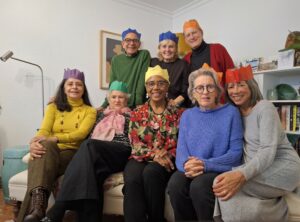Five women sitting on a couch and behind them a woman flanked by two men standing. Each is wearing a paper crown.