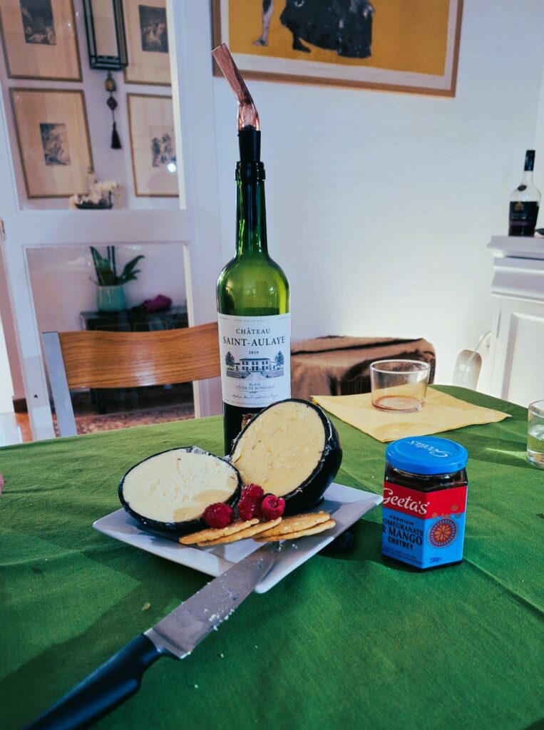 A siced open ball of cheese encased in black wax sits on a square plate with several crackers topped by raspberres and a knife on the side. next to the plat is a jar of chutney and behind it a bottle of wine. Everything sits on a green tablecloth.