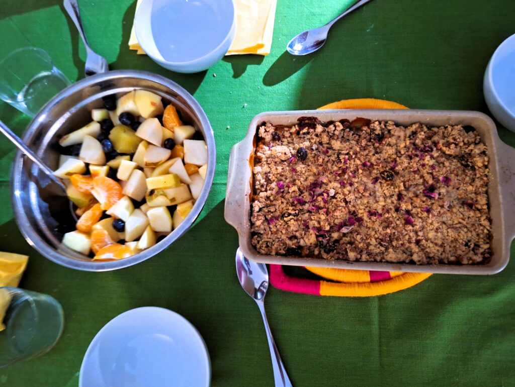 A stainless steel bowl of cut of fruit next to a rectangular pan of apple crisp sit on a green tablecloth with small white serving bowls and spoons at the top and the bottom of the photo.