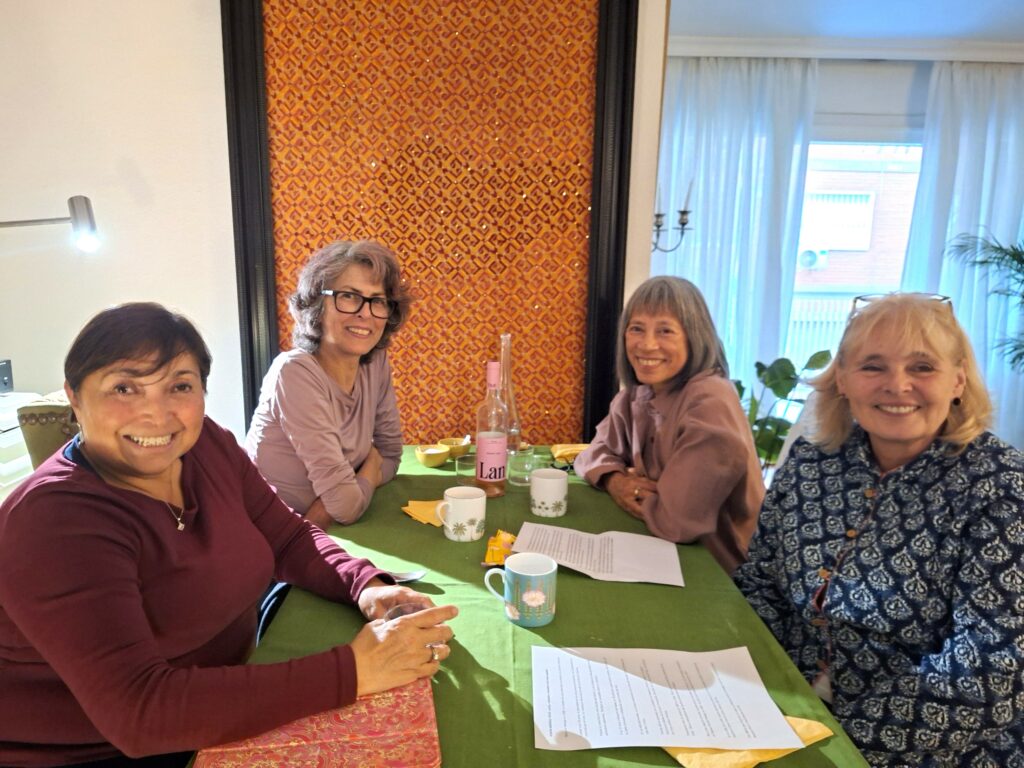 Four women at a diing room table on which sheets of paper are spread about and a bottle of wine. All the women are smiling and looking at the camera.