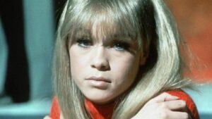Color photo of the young singer Marisol. Long blond hair with long bangs. She's unsmiling with a pensive, almost sad look. Her arms ae crossed to hug her shoulders.