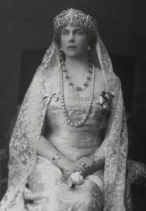 Black and white photo of Spanish Queen Victoria Eugenia whose crown holds a veil that cascades down her shoulders. She wears two necklaces as well as bracelets on each wrist. She is unsmiling and regal.
