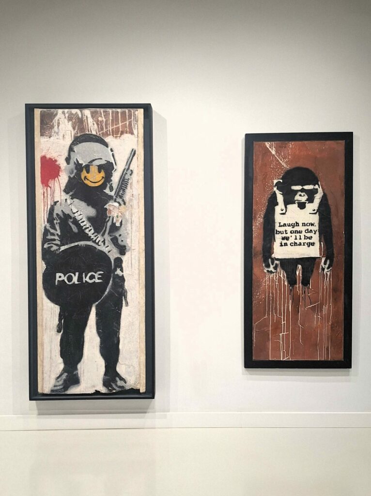 Photo of two Bansky paintings, one showig a policeman with a smiley face and the other showing a monkey with a sign that says, laugh now, but one day we'll be in charge.