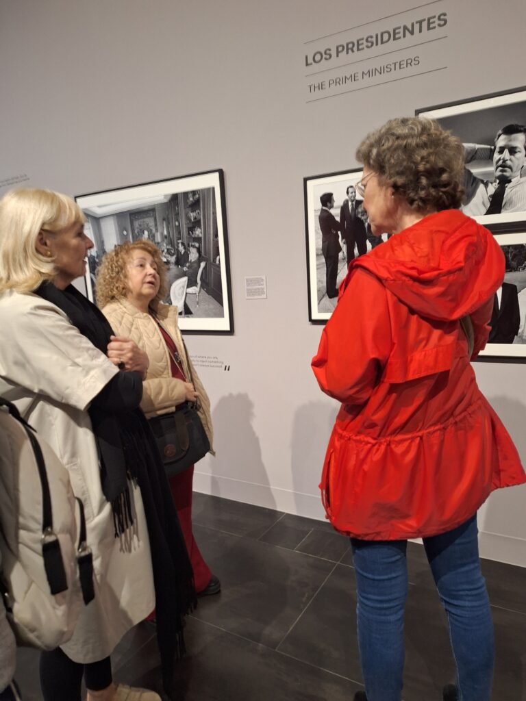 Three women have a discussion in front of a photo exhibit called Los Presidentes.