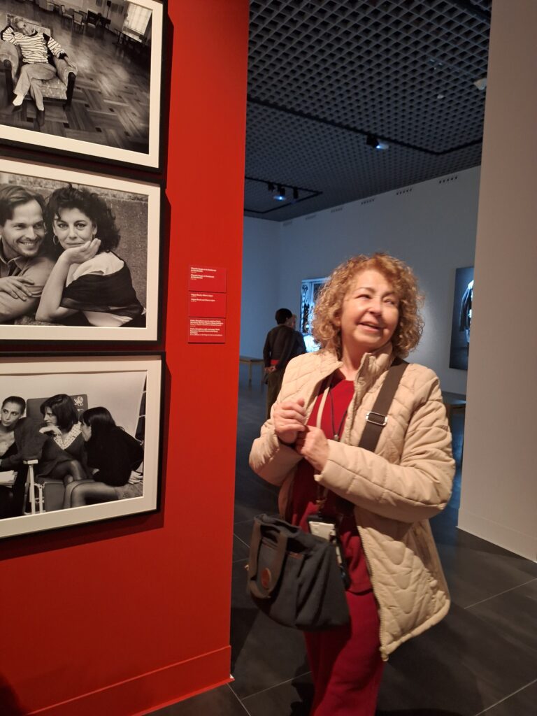 A woman with light brown hair and wearing a red top and beige jacket stands next to a series of black and white photos of celebrities of 70s and 80s Spain.