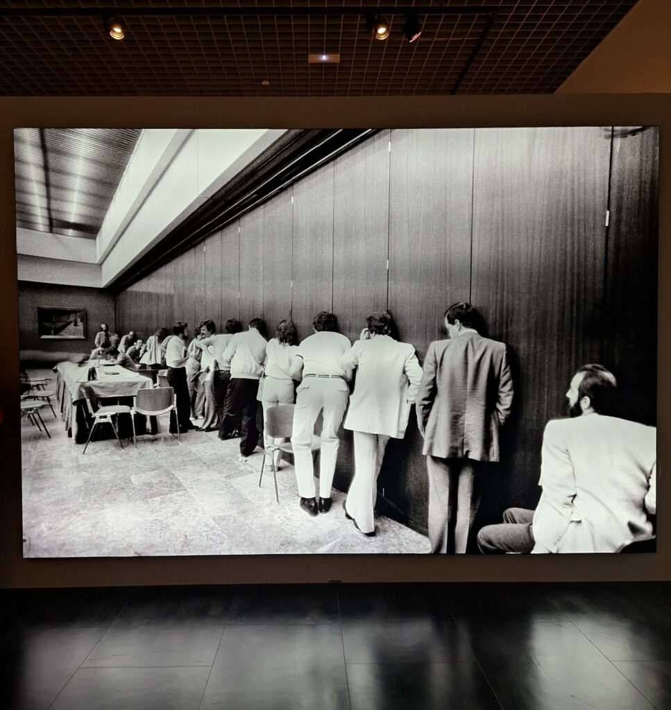 Black and white photo showing a line of journalists with their ears against a partition trying to hear the proceedings happenng in the next room.