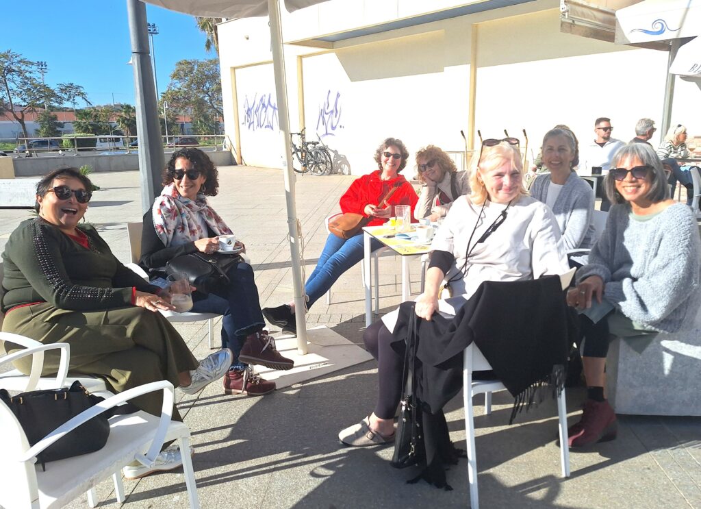 Six women are seated around a table of drinks outdoors on a sunny day.