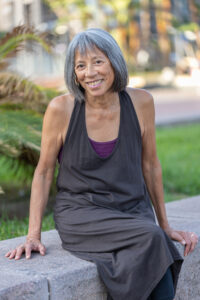 Brown-skinned woman with short graying hair and dressed in a sleeveless brown shift sits on a bench smiling.