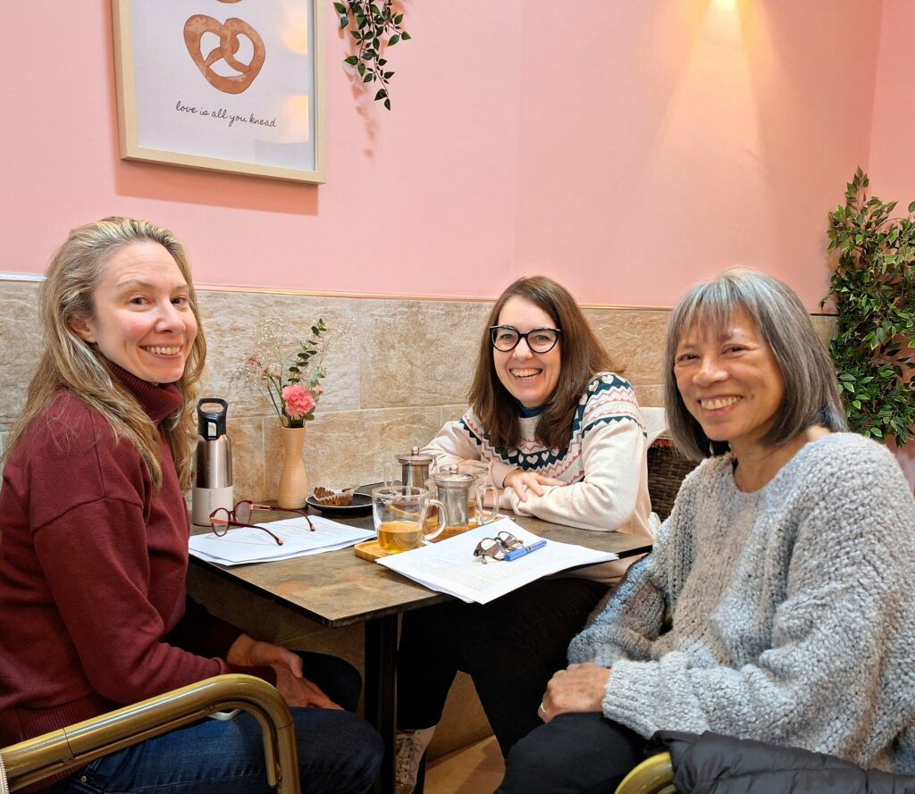 Three women sit at a small table in a coffee shop looking at the camera. There are papers and cups of tea on the table.