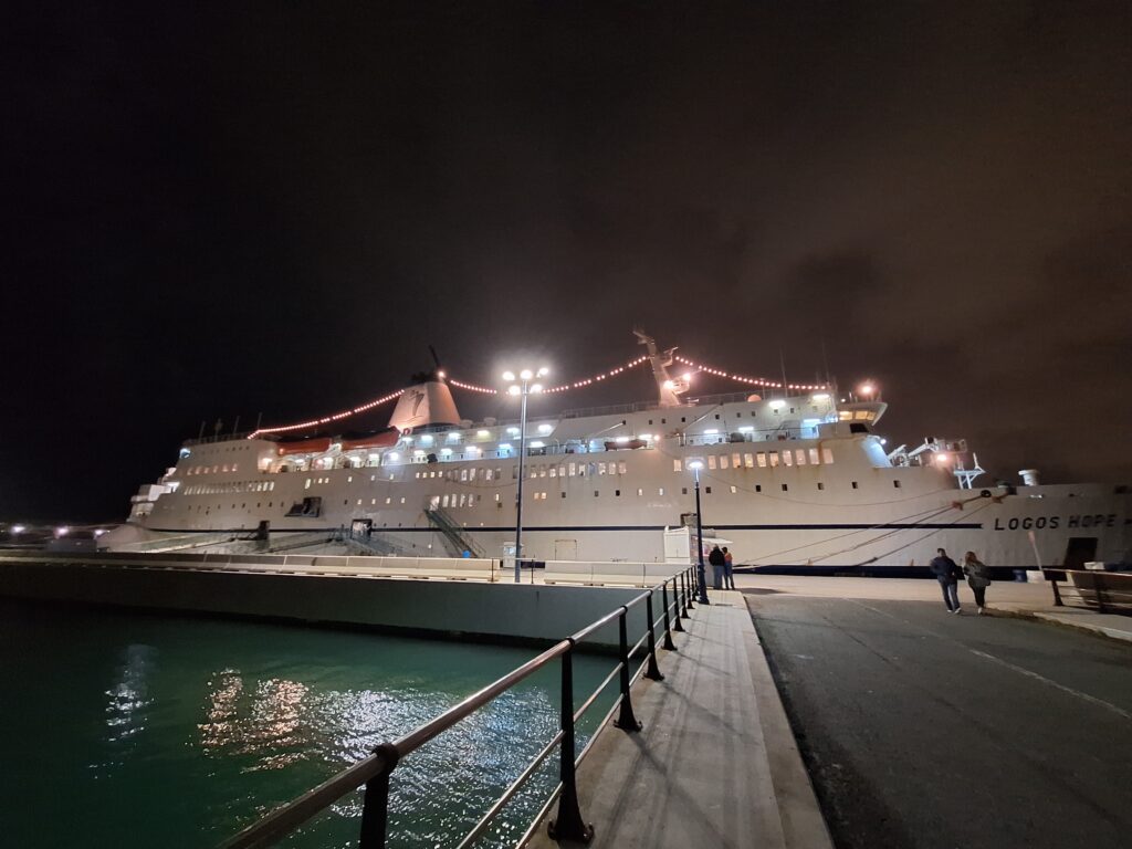 A docked ship at night with lights on with two couples walking toward it.