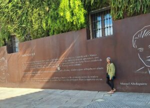 Woman in green sweater stands in front of a wall commemorating the founding of Imprenta Sur in Malaga