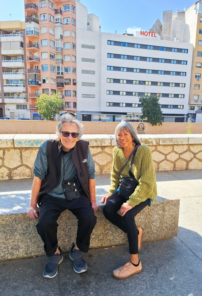 A man and awoman sit in a stone bench on a sunny day with a low brick wall and a series of multi-story buildings behind them.