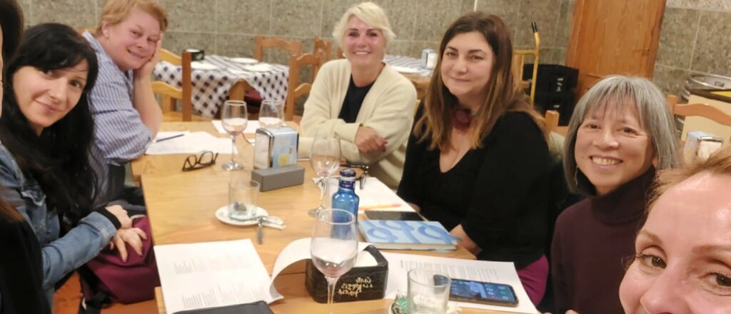 A group of five women of various nationalities sit around a restaurant table with papers in front of them.