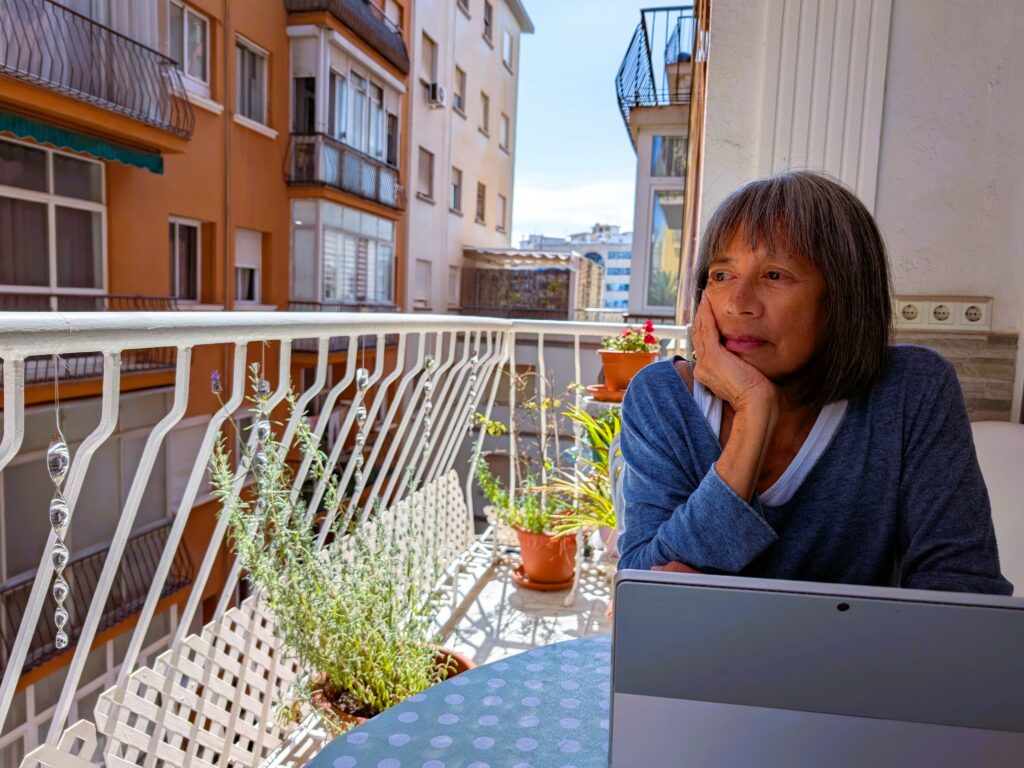 A woman with short dark haur and her chin cupped in her hand sits at a table on a balcony and stares into the dustance. The balcony rail is white and there are plants on the balcony.